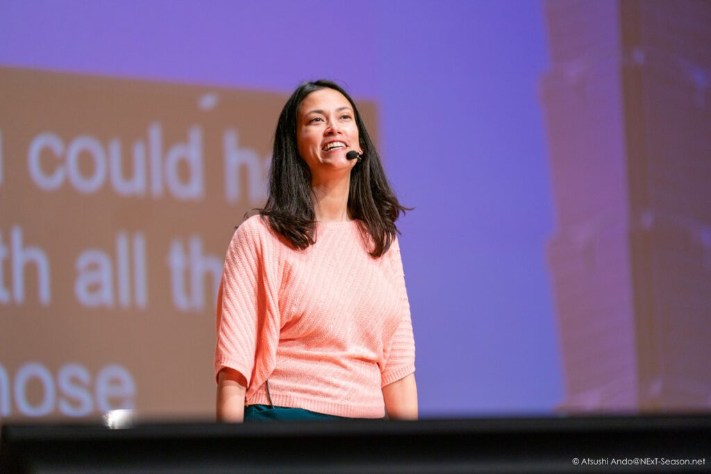 A woman is speaking on stage, wearing a light pink sweater and dark green pants. She has long, dark hair and is smiling, appearing engaged with her audience. In the background, there are large text projections, likely displaying her speech or key points. The setting suggests a conference or presentation environment.