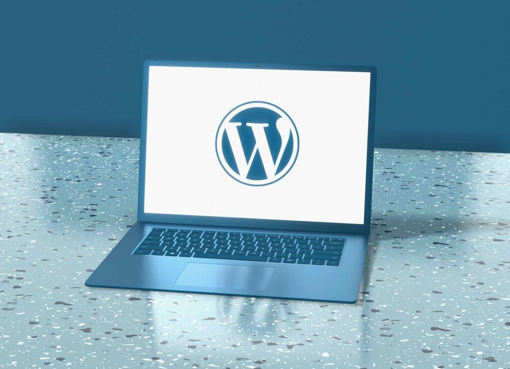 A laptop is open on a speckled surface, displaying the WordPress logo in the center of the screen. The laptop has a sleek design with a blue hue, and the background features a solid blue wall. The surface beneath the laptop is glossy with a mix of light and dark speckles.