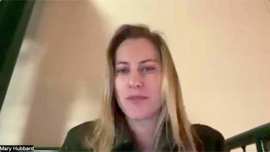 A woman with long, light brown hair is sitting in front of a neutral-colored background. She is wearing a dark jacket and appears to be engaged in conversation. The setting suggests an indoor environment, possibly during a video call, with a hint of a staircase visible in the lower part of the frame. The lighting is soft, creating a warm atmosphere.