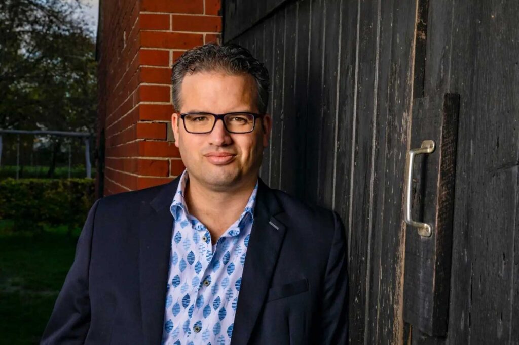 A man stands confidently against a rustic wooden wall, partially covered by a dark, weathered door. He wears a dark blazer over a light blue shirt with a pattern of stylized leaves. His hair is short and slightly tousled, and he sports glasses. The background features a blurred green landscape, suggesting an outdoor setting. The lighting is warm, highlighting his features.