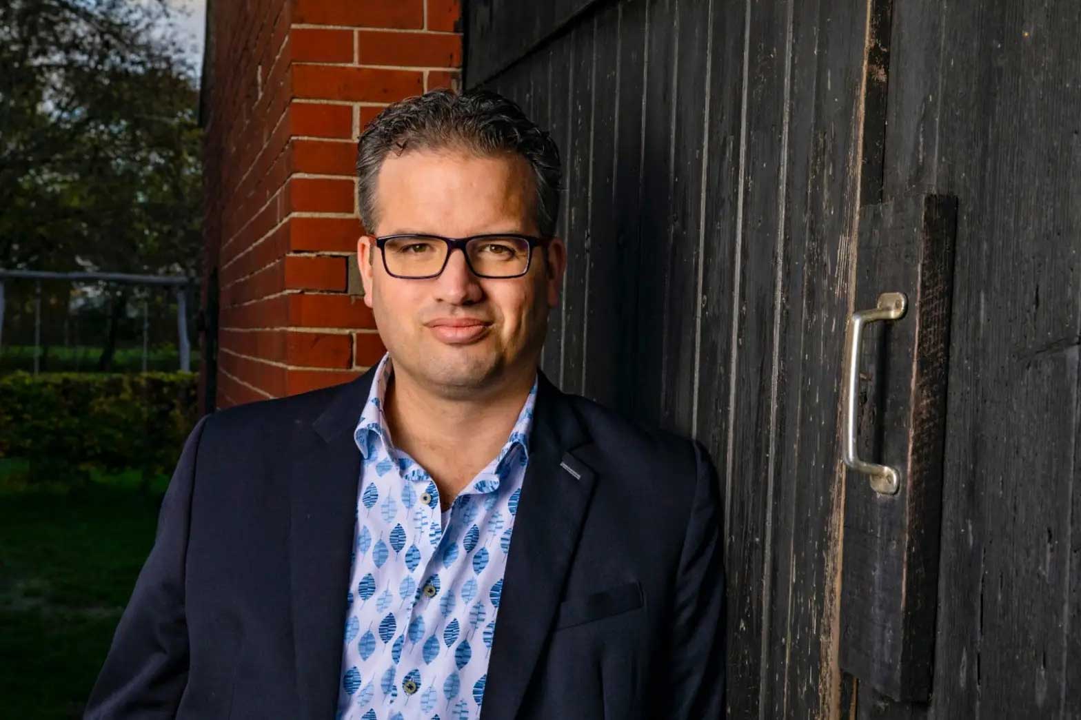 A man stands confidently against a rustic wooden wall, partially covered by a dark, weathered door. He wears a dark blazer over a light blue shirt with a pattern of stylized leaves. His hair is short and slightly tousled, and he sports glasses. The background features a blurred green landscape, suggesting an outdoor setting. The lighting is warm, highlighting his features.
