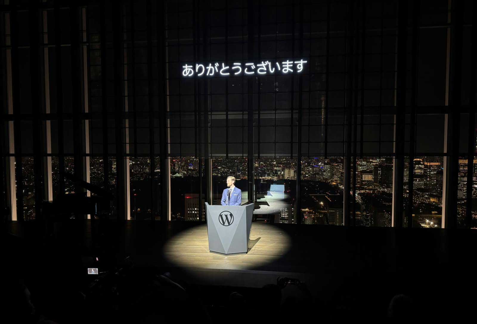 A speaker stands at a podium with a logo, illuminated by a spotlight. Behind them, large windows reveal a panoramic view of a city skyline at night, filled with lights. Above the speaker, text in Japanese reads "ありがとうございます," which translates to "Thank you." The setting appears to be a formal event or presentation.
