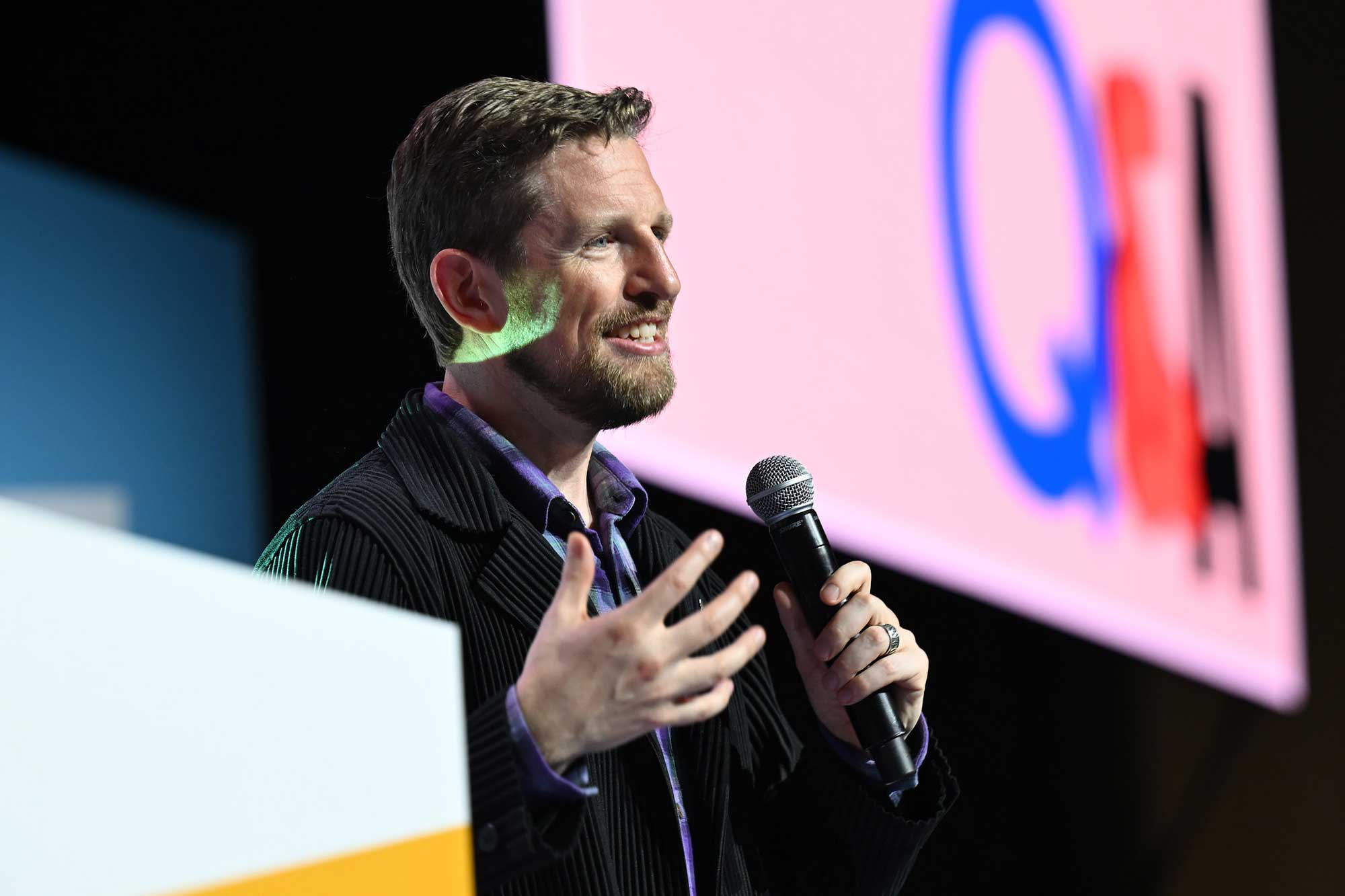 A man with a beard is speaking into a microphone, gesturing with his hands as he engages with an audience. He is wearing a black striped jacket over a purple shirt. In the background, a large screen displays the text "Q&A" in bold colors. The setting appears to be a conference or event stage.