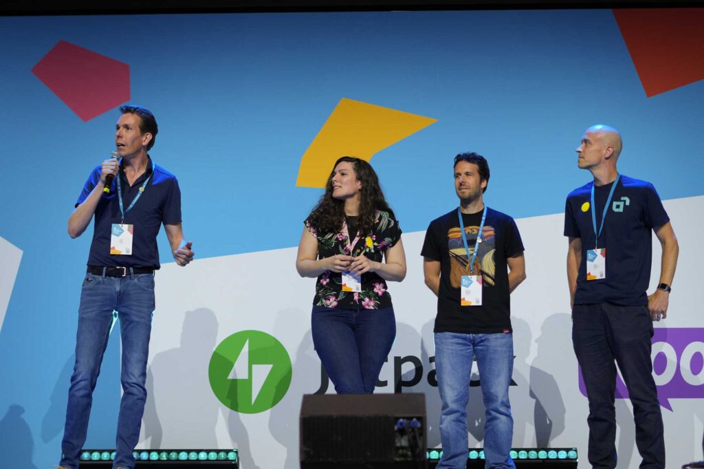 A group of four individuals stands on stage, engaged in a presentation. The first person, on the left, holds a microphone and is speaking. They wear a blue shirt and jeans. Next to them, a woman in a floral top appears to be listening attentively, holding something in her hands. The third person, wearing a black t-shirt with a graphic design, stands calmly, while the fourth person, on the right, is dressed in a dark shirt and looks towards the audience. The background features colorful geometric shapes and logos, including a green logo with arrows and text that reads "Jetpack" and "Koo.