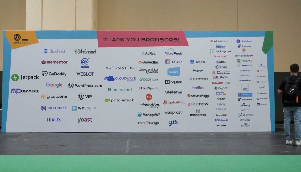 A large banner displays various logos of sponsors for the WCEU 2024 event. The top section features a "Thank You Sponsors!" message in bold letters. Below this, logos from companies like Bluehost, Elementor, Jetpack, Google, WooCommerce, and many others are arranged in a grid format. The background is colorful, with sections in blue, pink, and green, and a person stands to the right, facing the banner. The floor is covered in green material.