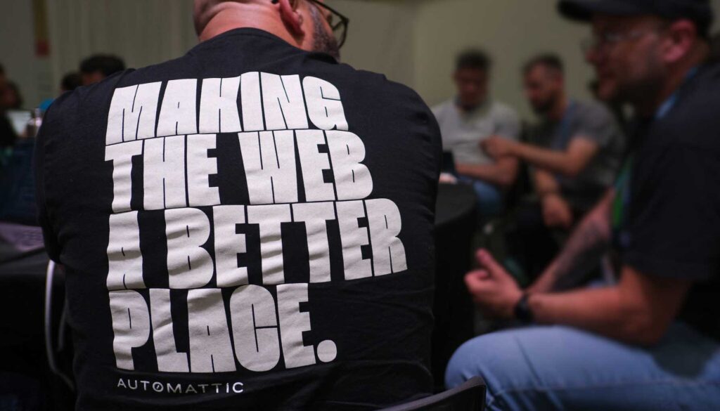 A person is seated with their back to the camera, wearing a black t-shirt that features bold white text reading "MAKING THE WEB A BETTER PLACE." Below the text, the word "AUTOMATTIC" is printed in smaller letters. In the background, several individuals are engaged in conversation, seated around a table in a casual setting.