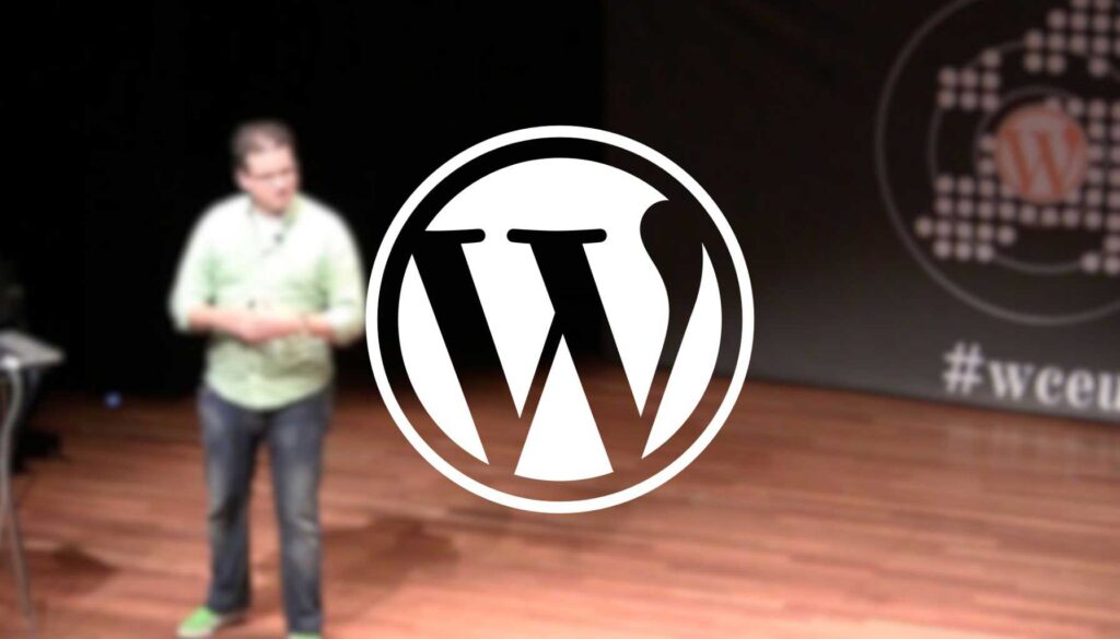 A speaker stands on a wooden stage, engaging with the audience. The speaker is wearing a light green shirt and appears to be gesturing as they talk. In the background, there is a large banner featuring the WordPress logo, which is prominently displayed in the center of the image. The setting suggests a conference or presentation related to WordPress, indicated by the hashtag "#wceu" visible on the banner.