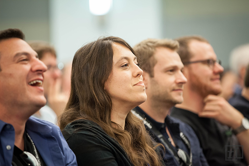 A group of people is seated in an audience, engaged and smiling. The focus is on a woman with long, dark hair, who appears thoughtful. To her left, a man is laughing, while another man next to her has a neutral expression. The background shows a blurred audience, indicating a lively event or presentation. The lighting is bright, contributing to a positive atmosphere.
