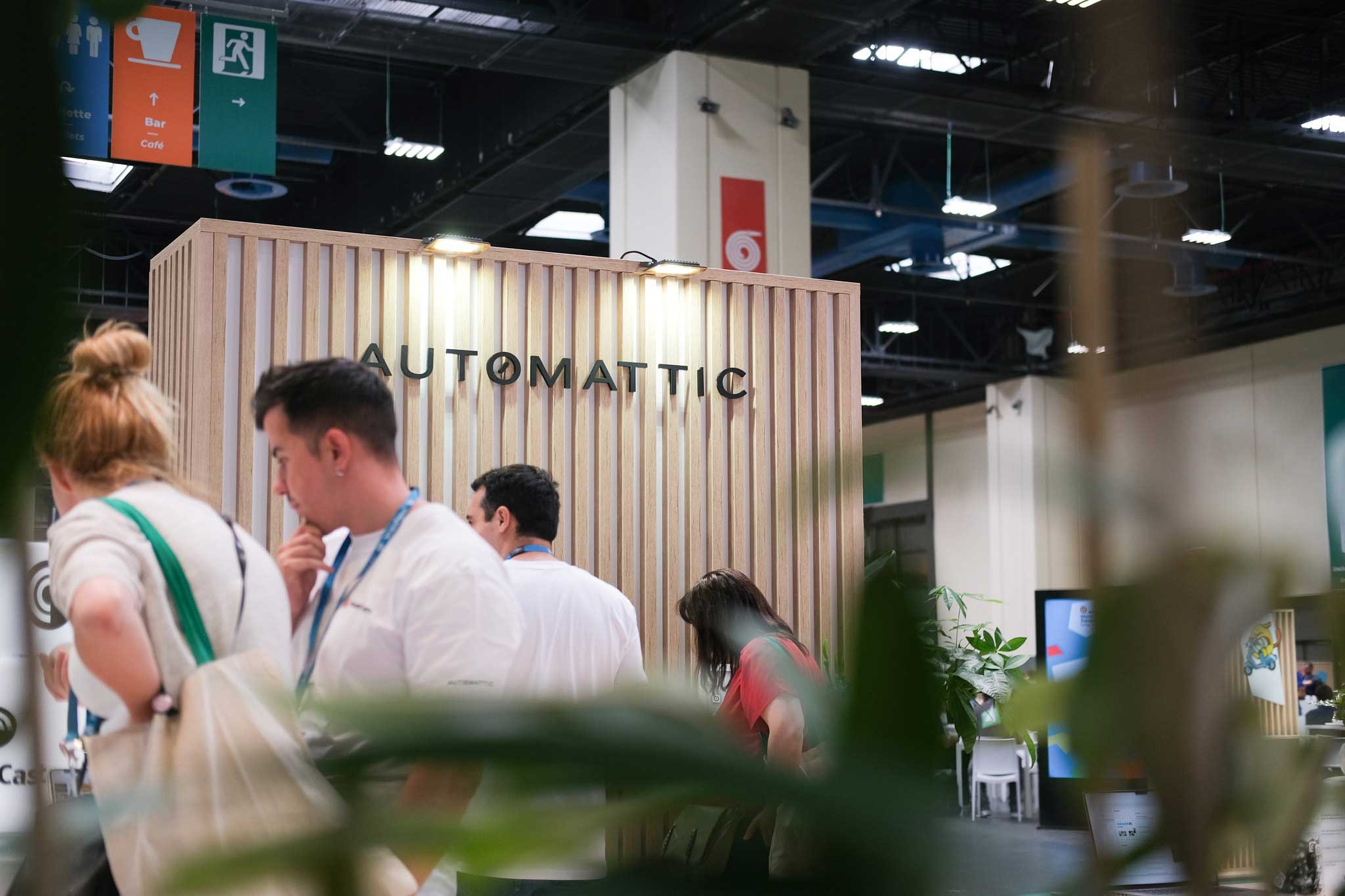 A wooden booth with vertical slats features the name "AUTOMATTIC" prominently displayed. In the foreground, several people are engaged in conversation, with one person in a white shirt thoughtfully touching their chin. The background includes signs for amenities like a café and bar, along with some greenery partially obscuring the view. The setting appears to be a conference or exhibition space.