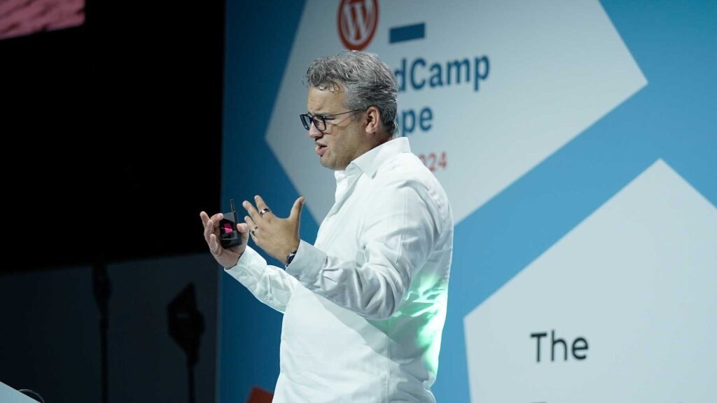 A speaker is presenting on stage, gesturing with both hands while holding a device in one hand. They are wearing a white shirt and glasses. The background features a blue and white design with the WordCamp logo and text indicating the event is for 2024. The speaker appears engaged and animated, emphasizing their points during the presentation.