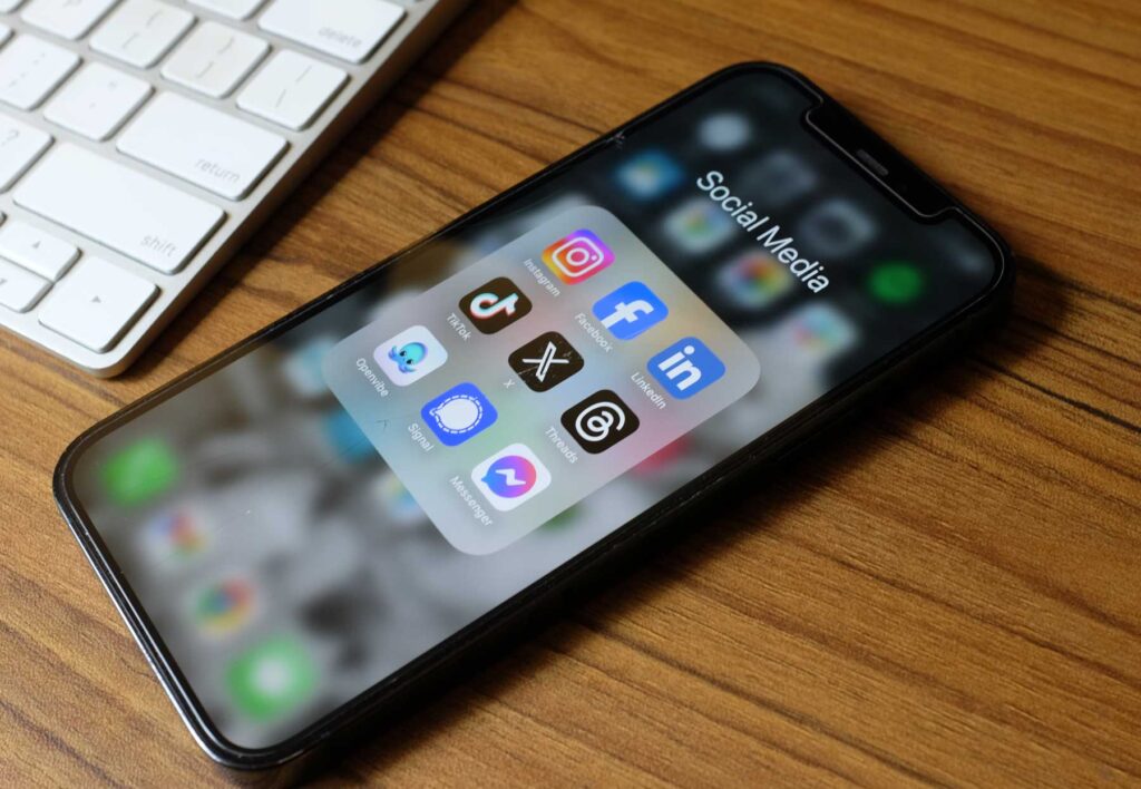 A smartphone is displayed on a wooden surface, showing a folder labeled "Social Media" on its screen. Inside the folder are icons for various social media apps, including Instagram, Facebook, TikTok, LinkedIn, and others. A white keyboard is partially visible in the background. The phone has a sleek design with a black border.