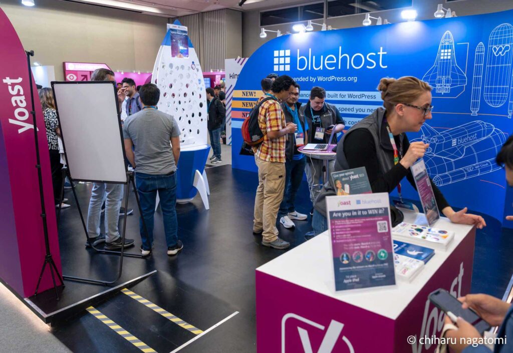 A busy exhibition hall at a WordPress event featuring booths from Bluehost and Yoast. Attendees engage in discussions and explore promotional materials. The Bluehost booth has a blue backdrop with technical illustrations, while the Yoast booth features a magenta display with a contest sign-up station.