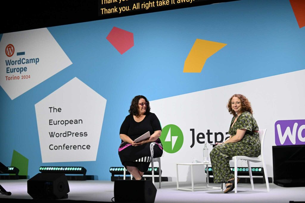 Two women are seated on stage at a conference, engaging in conversation. One woman, wearing a black top and a patterned skirt, holds a document. The other woman, dressed in a green patterned jumpsuit, sits beside her. The backdrop features colorful geometric shapes and text promoting "WordCamp Europe 2024" in Torino, along with logos for Jetpack and WordPress. A glass of water and a small table are positioned between them. The setting appears to be a professional conference environment.