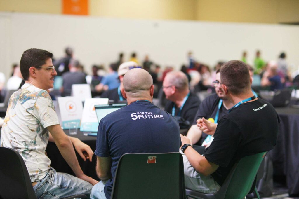 A group of people engaged in discussion at WordCamp Europe 2024. One person is wearing a navy blue t-shirt with '5 for the Future' printed on the back. The background shows a large room filled with attendees working on laptops and collaborating at tables.