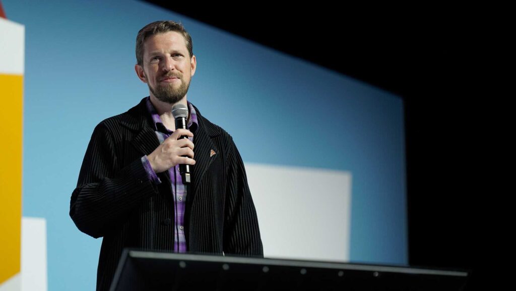 A man with a beard speaks into a microphone on stage, wearing a black jacket.