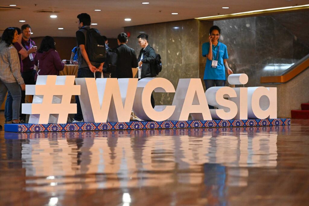 Large white letters spell out "#WCAsia" in a conference setting with attendees nearby.